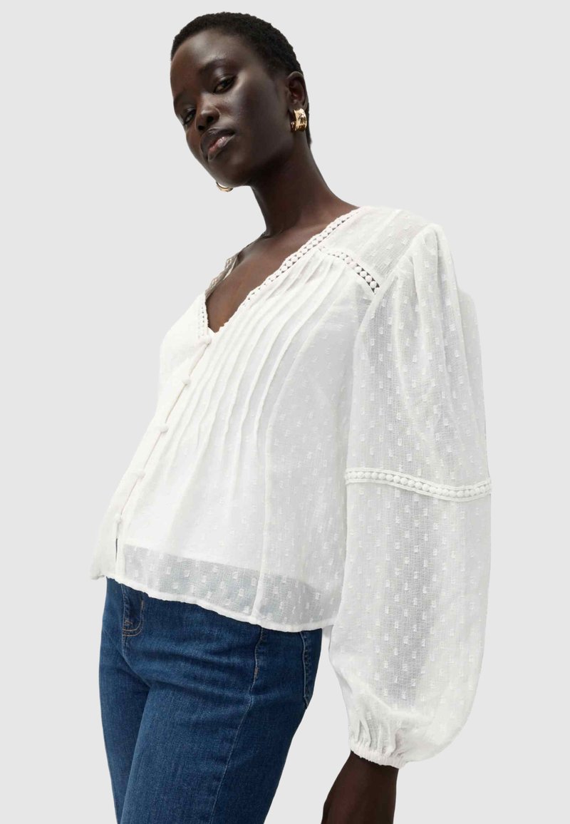Blusa bianca leggera con scollatura a V, chiusura con bottoni, tessuto texturizzato, dettagli plissettati e maniche a sbuffo voluminose.