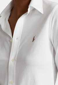 Chemise blanche boutonnée portée par une personne à la peau foncée, avec un petit logo brodé d'un joueur de polo sur le côté gauche de la poitrine.