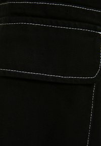 Mustan farkkukankaan, jossa on kontrastivalkoinen tikkaus, näkyvissä oleva taskudetaili, joka esittelee sileää pintaa ja jämäkkää muotoilua.