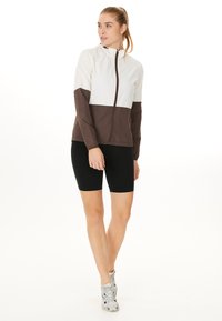 Lichte zip-up jas in crème en bruin, met een capuchon en lange mouwen. Gecombineerd met zwarte bike shorts en grijze sneakers. Eenvoudig ontwerp.
