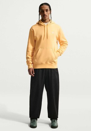 Mand med fletninger iført en gul Nike-hættetrøje, løse sorte bukser og grønne sneakers, står med den ene hånd i lommen.