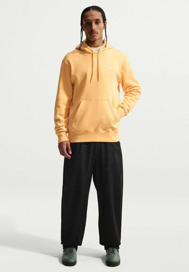 Mann mit geflochtenem Haar, der einen gelben Nike-Hoodie, lockere schwarze Hose und grüne Sneaker trägt, steht mit einer Hand in der Tasche.