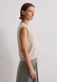 Beiges, ärmelloses Strickoberteil mit strukturiertem Muster und durchgehender Knopfleiste vorne. Kombiniert mit einem grauen, plissierten Rock.