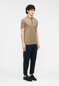 Καφέ πόλο μπλουζάκι με γιακά και φερμουάρ, συνδυασμένο με ναυτικά παντελόνια φόρμας με ρίγες στο πλάι. Το μοντέλο φοράει μαύρα αθλητικά παπούτσια. Μινιμαλιστικός σχεδιασμός.