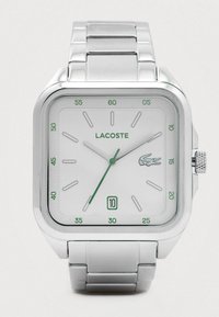 Sudraba Lacoste rokas pulkstenis ar kvadrātveida seju, baltu ciparnīcu, zaļiem akcentiem, datuma rādītāju 6 pulkstenī un metāla saiti.