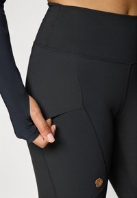 Hoge taille zwarte leggings met een gladde textuur, voorzien van zijzakken en een klein bruin logo detail op de dij.