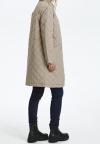 Beige quiltet frakke med en lang, afslappet pasform, sidelommer og knaplukning. Båret over mørke skinny jeans og kraftige sorte støvler.