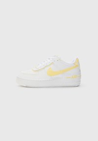 Bijela tenisica s žutim Nike logom, glatkim kožnim materijalom, zaobljenim vrhom i teksturiranom gumenom potplatom s "AIR" detaljem na peti.
