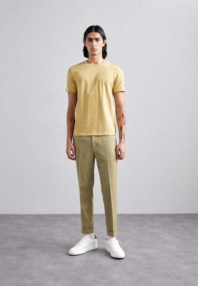 Camiseta mostaza combinada con pantalones verde oliva claro. El modelo lleva zapatillas blancas y está de pie sobre una superficie gris texturizada.