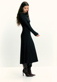Fekete hosszú ujjú ruha, testhez simuló szabás, puha anyag, midi hosszúságú, térdig érő bordó magas sarkú csizmával párosítva. Sima textúra, mintázat nélkül.