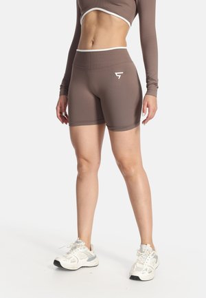 Žena nosiaca hnedé športové šortky s vysokým pásom a crop top s dlhým rukávom a biele tenisky, stojaca na jednoliatom pozadí.