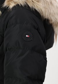 Zwarte pufferjas met een kraag van imitatiebont. Heeft een ronde zwarte patch met het merklogo en een doorgestikte textuur op de stof.