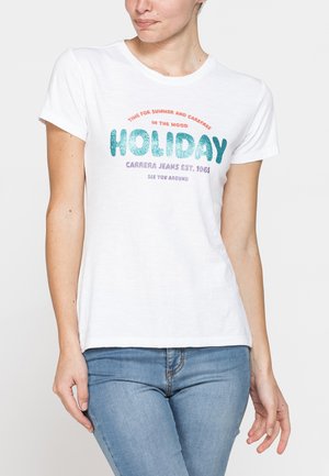 Donna che indossa una maglietta bianca con testo colorato "HOLIDAY" e jeans blu, in piedi davanti a uno sfondo chiaro e uniforme.