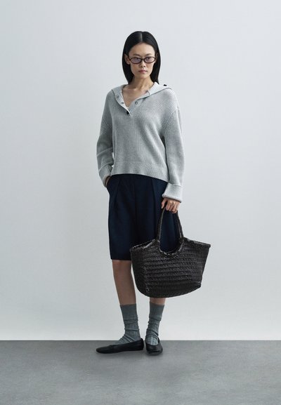 Γκρι πλεκτό πουλόβερ με κουκούλα και λεπτομέρεια κουμπιών, μπλε σορτς, υφασμάτινη σκούρα τσάντα, γκρι κάλτσες και μαύρα επίπεδα παπούτσια μπροστά σε ουδέτερο φόντο.