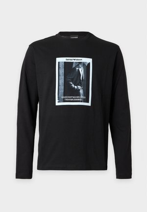 Jeune homme portant un t-shirt à manches longues noir avec un motif graphique, un jean bleu et des baskets noires, debout devant un fond blanc uni.