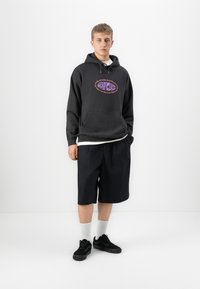 Jonge man in een zwarte hoodie met grafische tekst, zwarte knielange shorts, witte sokken en zwarte sneakers die tegen een plain witte achtergrond staat.