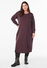 Žena s dugom smeđom kosom u bordo midi haljini dugih rukava s bočnim prorezom, crnim tajicama i tamnim gležnjačama, stoji i smiješi se.