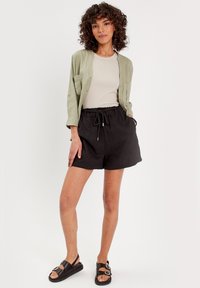 Chemise boutonnée vert clair sur un top beige, associée à un short noir ample et des sandales noires avec des accents de boucle. Textures lisses visibles.