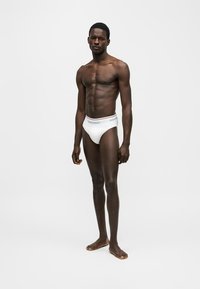 Ευρύς, μελαψός άνδρας στέκεται ξυπόλυτος φορώντας λευκά εσώρουχα Dsquared2 μπροστά σε ένα απλό ανοιχτό γκρι φόντο.