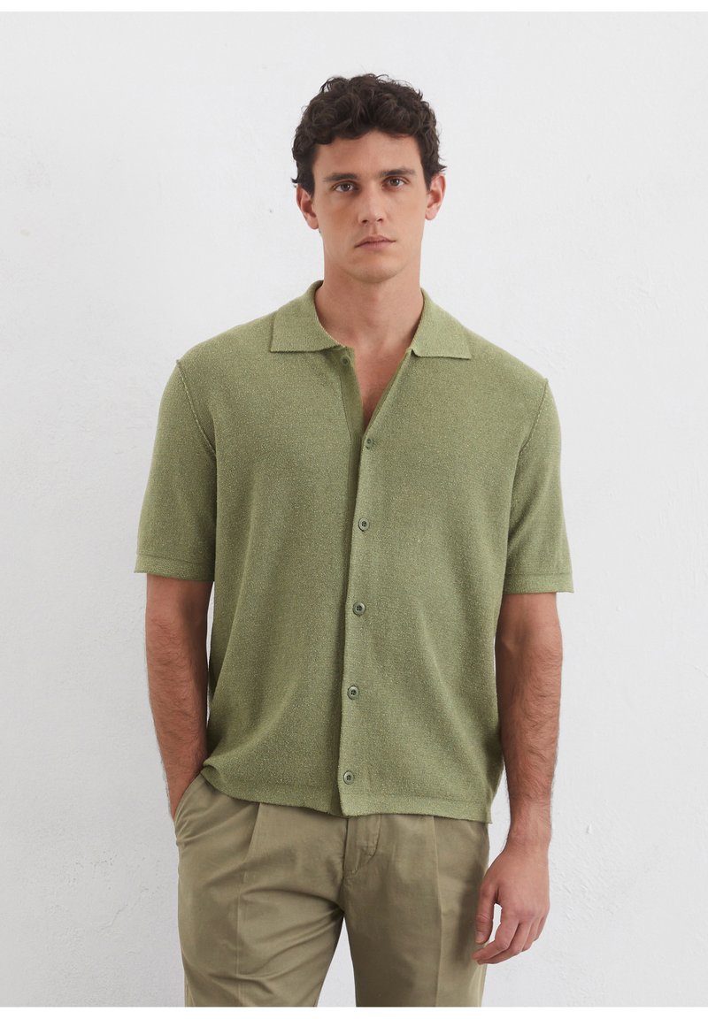 Chemise vert clair à manches courtes avec col, en tissu texturé et cinq boutons sur le devant. Associée à un pantalon beige.