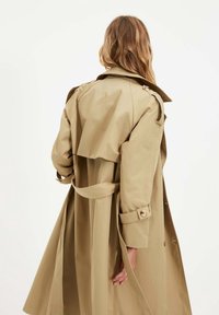 Béžový trenčkot s páskem v pase, dvojím zapínáním na knoflíky, nášivkami na ramenou a volným střihem. Měkká látka s hladkou texturou.