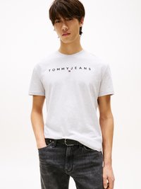 Junger Mann mit dunklen Haaren, der ein hellgraues "Tommy Jeans"-T-Shirt und dunkle Jeans trägt, mit einer Hand in der Tasche.
