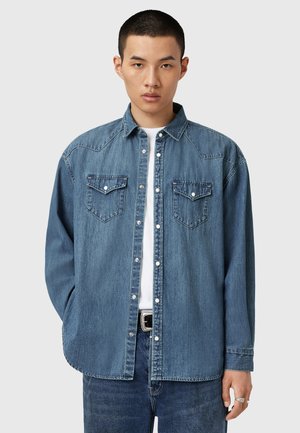Denim shirt in medium blue, featuring snap buttons, two chest pockets, pointed yoke detail, and long sleeves with buttoned cuffs.