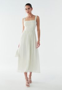 Gaiša, teksturēta midi kleita mīkstā piena krāsā ar kvadrātveida izgriezumu, mežģīņu apdari un regulējamām lencēm. Valkāta kopā ar baltiem, siksniņām apaviem ar papēdi.