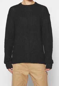 Czarny sweter z dzianiny z okrągłym dekoltem, prążkowaną strukturą i wydłużonymi rękawami. Nosi się go na jasnobeżowe spodnie.