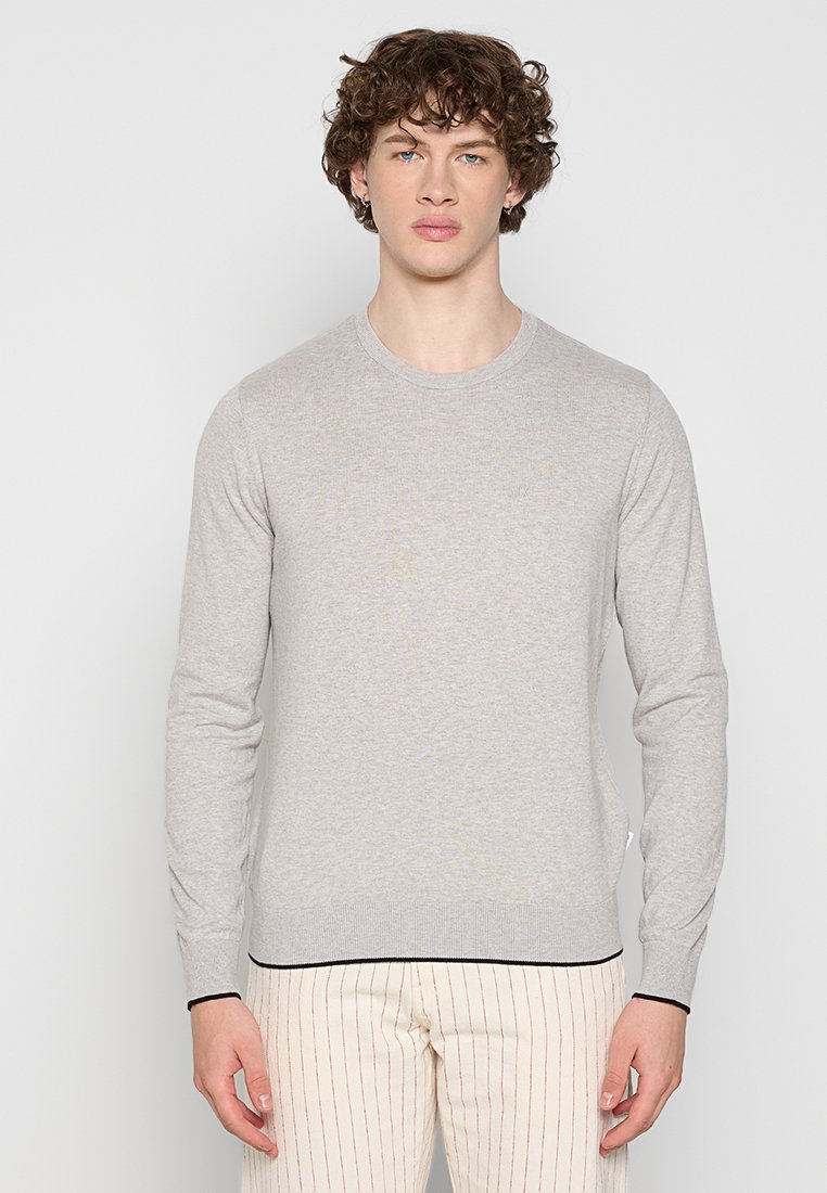 Junger Mann mit lockigem braunem Haar, der ein hellgraues Langarmshirt und cremefarbene Hose mit dünnen roten vertikalen Streifen trägt, vor einem weißen Hintergrund stehend.