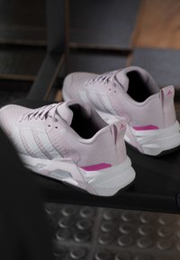 Ζευγάρι ανοιχτόχρωμα ροζ παπούτσια Adidas με λευκές σόλες και ρίγες τοποθετημένα σε ένα μαύρο πάγκο γυμναστηρίου.