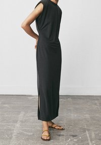 Person står i en ärmlös svart maxiklänning med sidofickor och bruna remsandaler på en betonggolv.