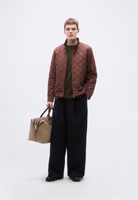 Καφέ μπουφάν με καπιτονέ σχέδιο, πράσινο τοπ, μαύρο φαρδύ παντελόνι, κρατώντας μια μπεζ και καφέ τσάντα τύπου ντουφελ με φερμουάρ και ανθεκτικές λαβές.