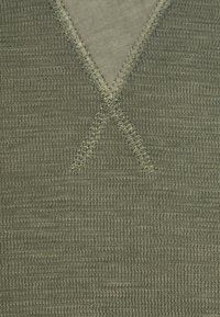 Nahaufnahme eines olivgrünen Strickstoffs mit verstärkten Nähten, die eine "X"-Form in der Nähe eines V-förmigen Rippenhalsauschnitts bilden.