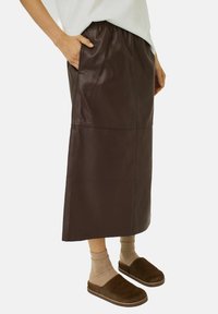 Jupe en simili cuir marron avec une taille élastique, des poches latérales et une coupe droite. Associée à des chaussettes marron jusqu'au genou et des pantoufles marron.