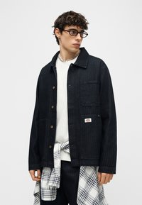 Νέος άνδρας με κοντά σγουρά μαλλιά και γυαλιά φοράει ένα σκούρο τζιν μπουφάν πάνω από ένα λευκό πουκάμισο, με ένα καρό πουκάμισο δεμένο γύρω από τη μέση.