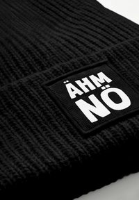 Black knitted beanie featuring a textured pattern. It has a rectangular patch with the text "ÄHM NÖ" in bold white letters.