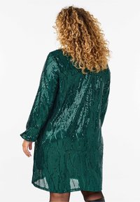 Grön paljettklänning med långa ärmar, med en flödande design och dekorativa mönster. Tyget har en glänsande, texturerad yta.