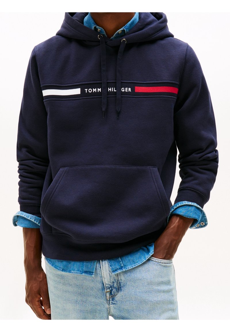 Granatowa bluza z kapturem z kieszenią z przodu, z białymi i czerwonymi poziomymi paskami oraz napisem „TOMMY HILFIGER”. Nałożona na koszulę jeansową.