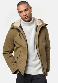 Giacca isolante color sabbia con cappuccio staccabile, colletto foderato in pile, tasche frontali e zip nere. Indossata sopra un maglione di lana bianco.