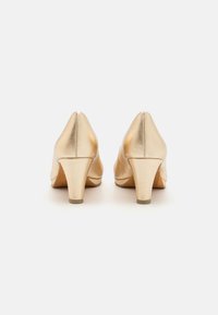 Chaussures à talons hauts en cuir doré avec un bout pointu et un talon bloc modéré. La texture est lisse avec une finition brillante et des détails de couture visibles.