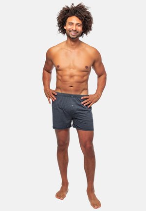 Pantaloni shorts grigi in cotone con elastico in vita, bottone frontale e vestibilità rilassata, indossati da un modello maschile senza camicia che sta in piedi a piedi nudi.