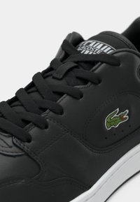Baskets en cuir noir avec lacets noirs, semelle blanche, écusson du logo Lacoste sur le côté et étiquette de la marque sur la languette.