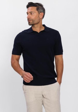 Man met kort donker haar en baard, gekleed in een zwart poloshirt met korte mouwen en licht beige broek, staand tegen een witte achtergrond.