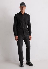 Mann, der ein schwarzes, langärmliges, Button-Up-Hemd, dunkelgraue Jeans, schwarze Schuhe und eine graue Baseballmütze trägt, steht an einer hellen Wand.