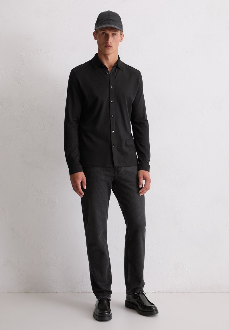Mann, der ein schwarzes, langärmliges, Button-Up-Hemd, dunkelgraue Jeans, schwarze Schuhe und eine graue Baseballmütze trägt, steht an einer hellen Wand.