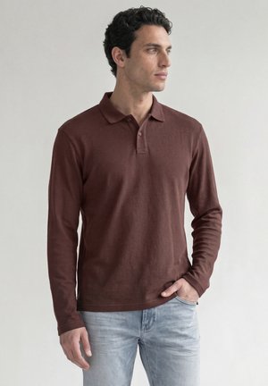 Homme portant un polo marron à manches longues et un jean bleu clair, debout avec une main dans la poche, regardant sur le côté.
