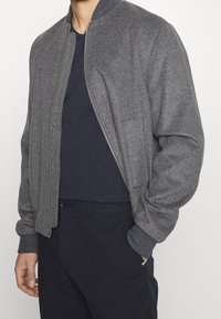Šedá bomber bunda se zipem vpředu, kulatým límcem a bočními kapsami. Vyrobena z texturované tkaniny, nošena přes námořnickou košili a tmavé kalhoty.