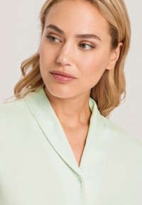 Ljusgrön blus med en mjuk och slät textur. Har en sjalkrage, knäppning med knappar och kontrasterande sömmar längs kanterna.