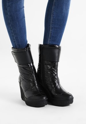 Bottines noires matelassées à semelle compensée avec fermetures éclair latérales, portées avec un jean slim bleu sur fond blanc.
